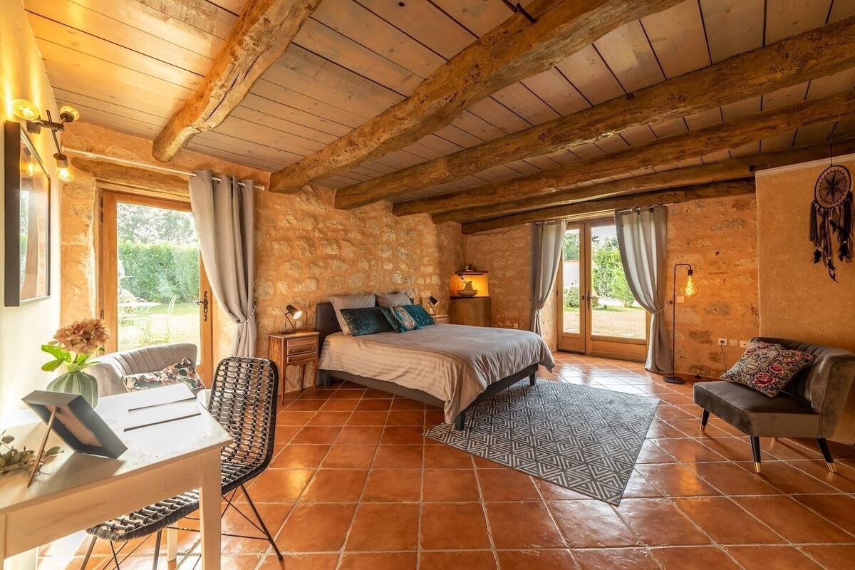 Une chambre à coucher au style rustique, avec un mur en pierre, un lit agrémenté de nombreux coussins, un bureau avec une chaise et une vue sur l’extérieur grâce à une grande fenêtre.