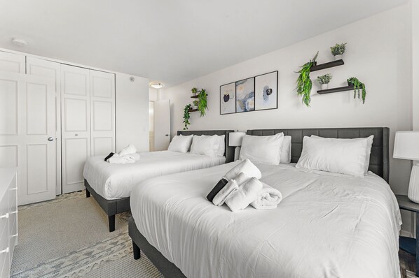 A hotel room with two beds, white bedding, a gray headboard, a bedside table with a lamp, and wall-mounted shelves with plants. - Gorgeous 2BR Next to BU f51x (Boston)