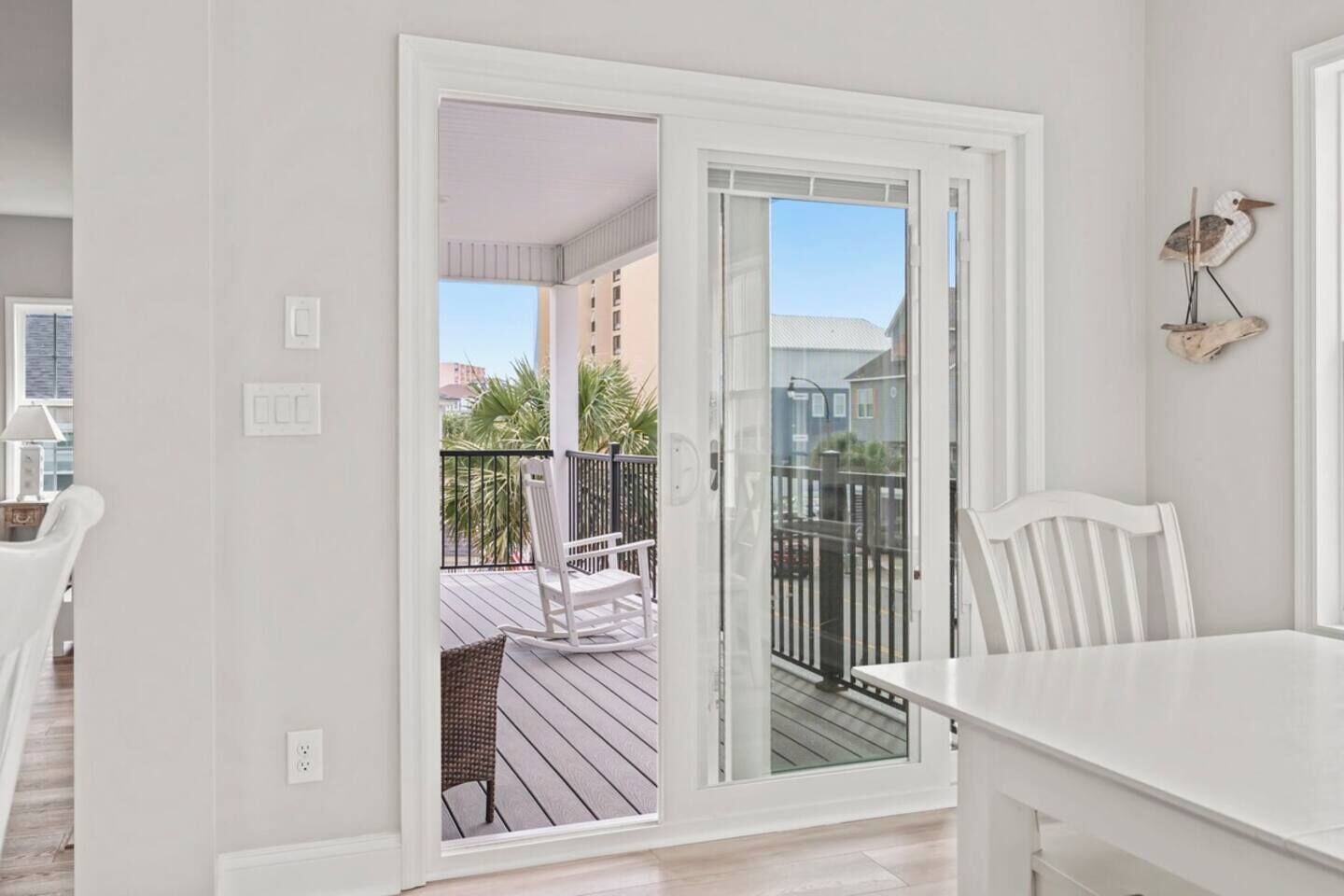 Une pièce avec une table et des chaises blanches, un balcon avec une chaise blanche, et une vue sur des immeubles et des palmiers.