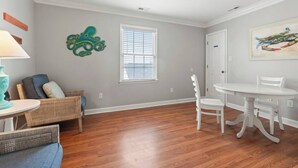 Une pièce comprenant une table à manger, des chaises, un canapé en osier, une fenêtre avec des stores et un élément décoratif fixé au mur.