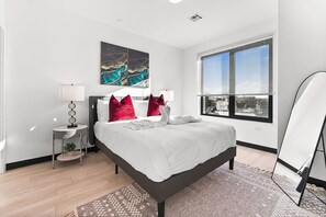 Une chambre moderne avec un lit, une table de chevet, une lampe, une grande fenêtre avec des stores et une vue sur la ville.