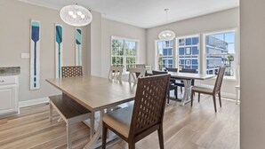 A dining area with a wooden table and chairs, pendant lighting, and large windows with a view of buildings. - Epic 7BR Getaway - Pool, Game Room, Put-put, Beach (North Myrtle Beach)