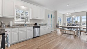 A modern kitchen with white cabinetry, a dishwasher, and a dining area with a table and chairs. - Epic 7BR Getaway - Pool, Game Room, Put-put, Beach (North Myrtle Beach)