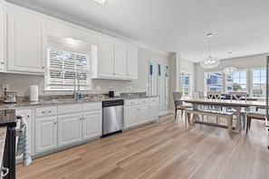 A modern kitchen with white cabinetry, a dishwasher, and a dining area with a table and chairs. - Epic 7BR Getaway - Pool, Game Room, Put-put, Beach (North Myrtle Beach)