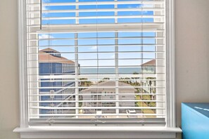 A window with horizontal blinds, a view of a building with a red roof, and a clear sky. - Epic 7BR Getaway - pool, game room, put-put, beach (North Myrtle Beach)