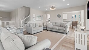 A spacious living room with a neutral color palette, a staircase, and a kitchen area visible through an open doorway. - Epic 7BR Getaway - Pool, Game Room, Put-put, Beach (North Myrtle Beach)