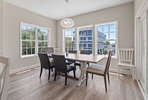 A dining area with a large table, four chairs, and a chandelier. There are large windows with a view of buildings outside.