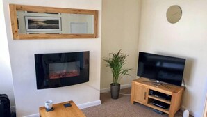 A living room with a fireplace, a television, a clock, and a coffee table. - Ringswell Gardens Room 3 Queen (Bath)
