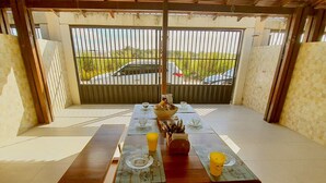 Uma área de refeições ao ar livre coberta, com uma mesa comprida posta para dois, bancos de cada lado e vista para uma estrada e vegetação através de portões de metal.