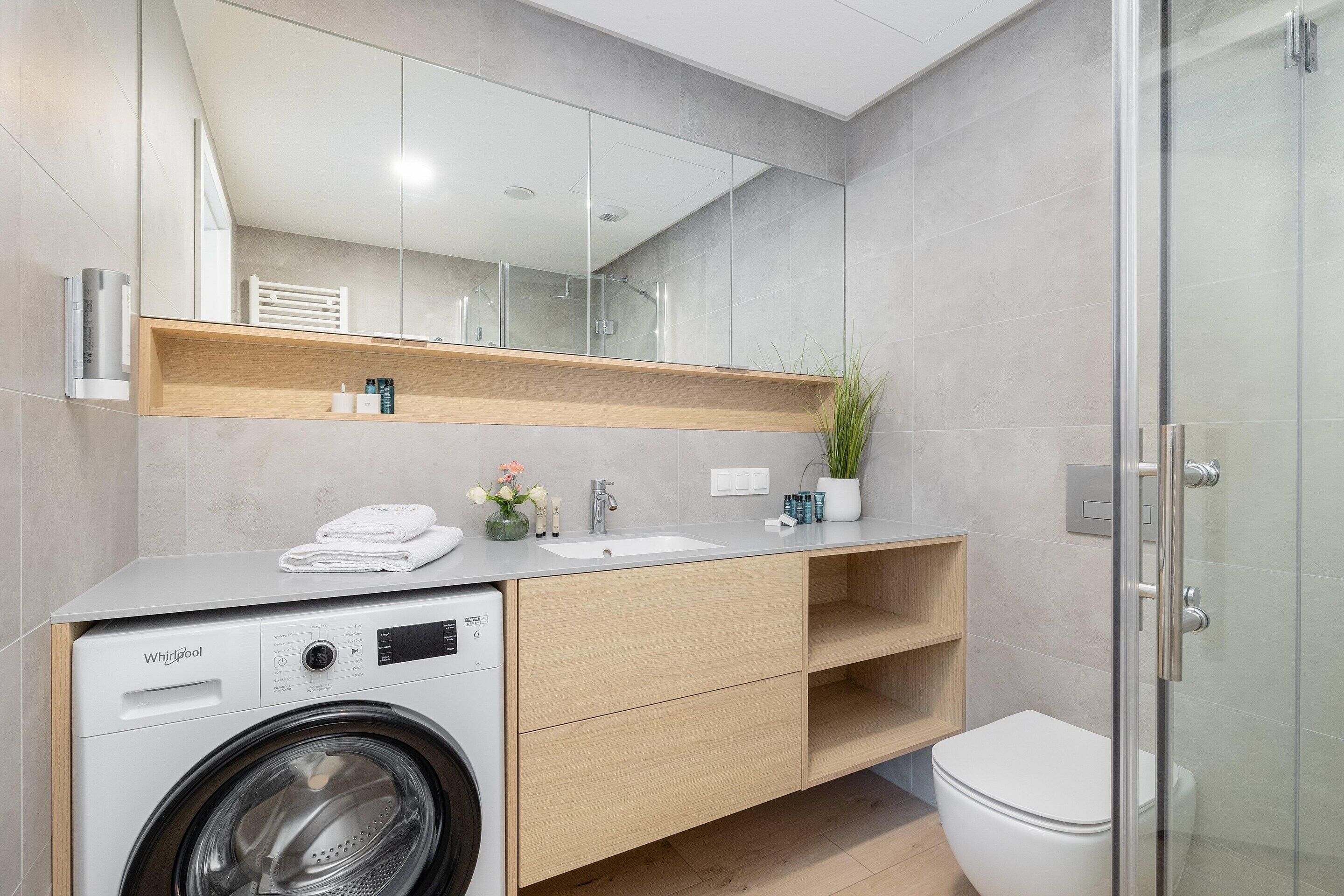 A modern bathroom with a washing machine, sink, mirror, and glass shower enclosure.