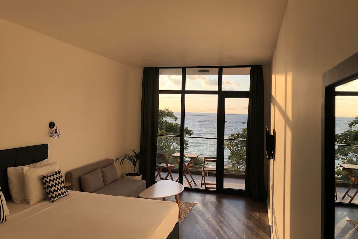 Un dormitorio con una cama grande, un sofá, una mesita y vista al mar a través de un ventanal amplio.