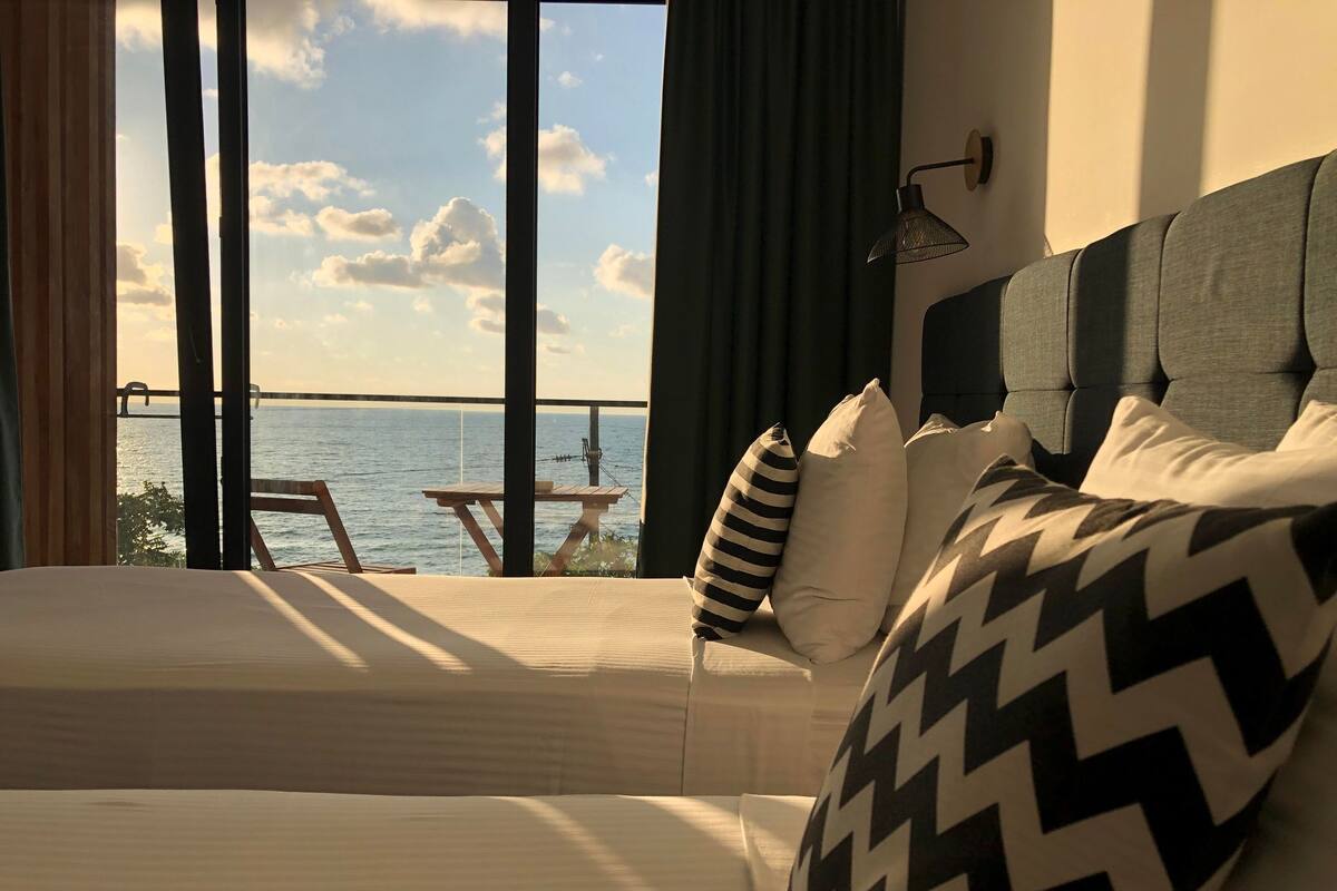 Habitación de hotel con dos camas, vista al mar, una mesa y sillas afuera, y un ventanal grande.