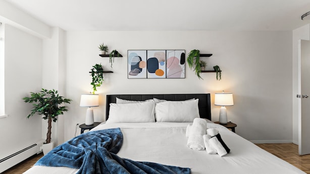 A hotel room with a large bed, two bedside lamps, a headboard, a wall-mounted abstract art piece, and potted plants. - Nice 2bd/1.5ba Next to BU 7qn5 (Boston)