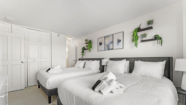 A hotel room with two beds, white bedding, a gray headboard, a bedside table with a lamp, and wall-mounted shelves with plants. - Beautiful 2bd/1.5ba Next to BU 74c3 (Boston)