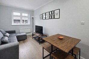 Uma sala de estar moderna com um sofá cinzento, uma mesa de jantar de madeira, uma televisão e quadros nas paredes.