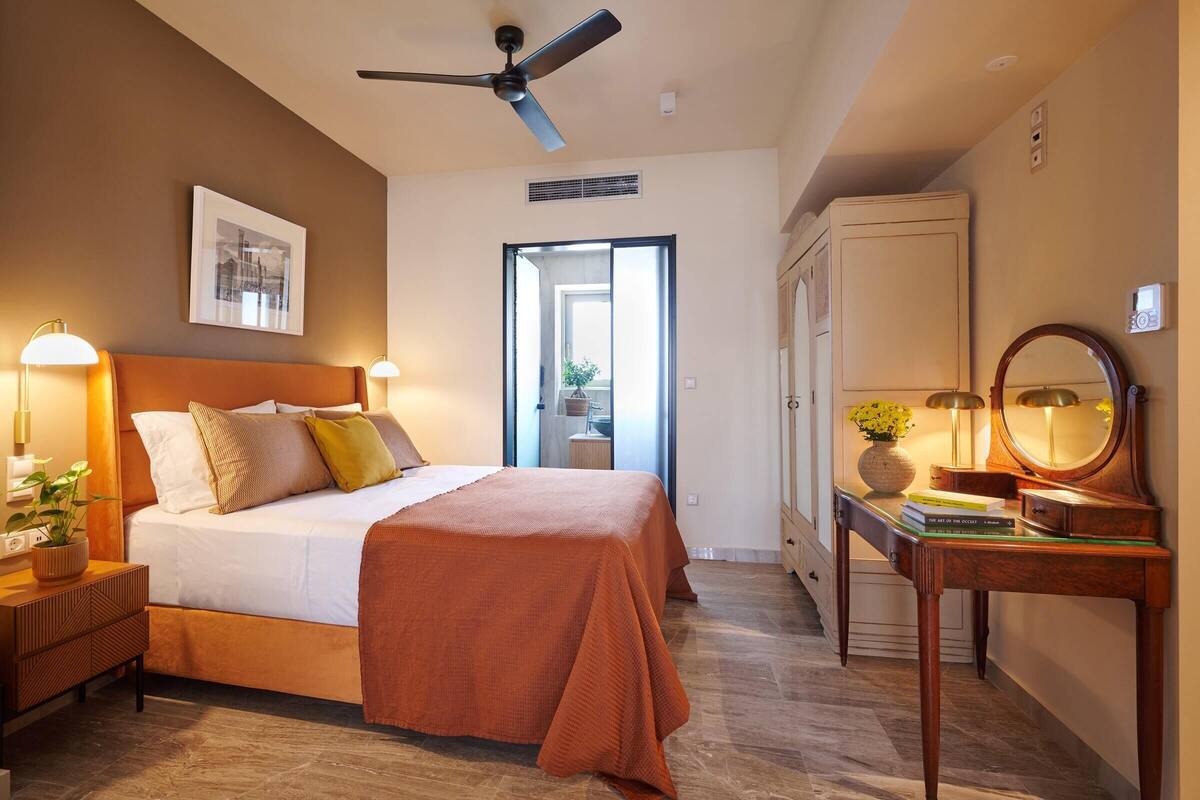 Ein Hotelzimmer mit Bett, Nachttischlampen, Deckenventilator, Schreibtisch mit Spiegel und Blick ins Freie.