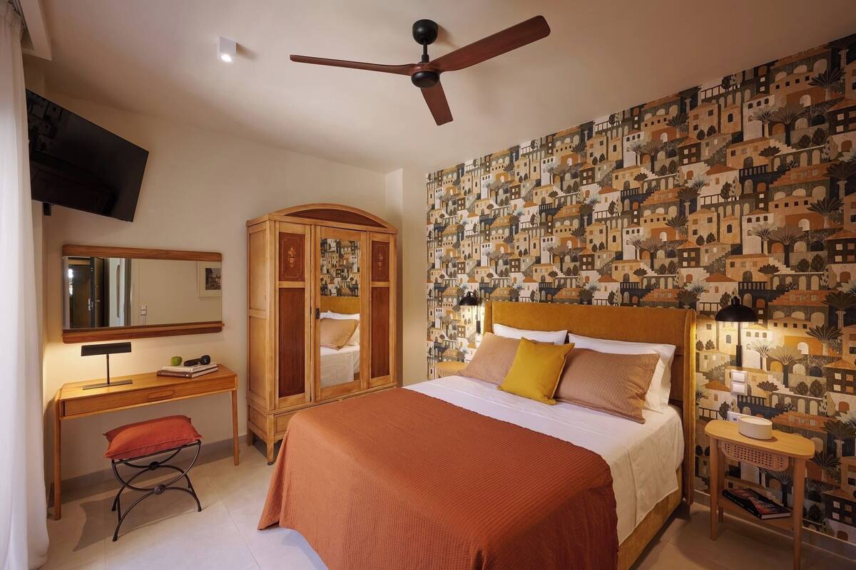 Ein Schlafzimmer mit Bett, Kleiderschrank, Schreibtisch, Stuhl, Deckenventilator und einer Wand mit Wandgemälde.