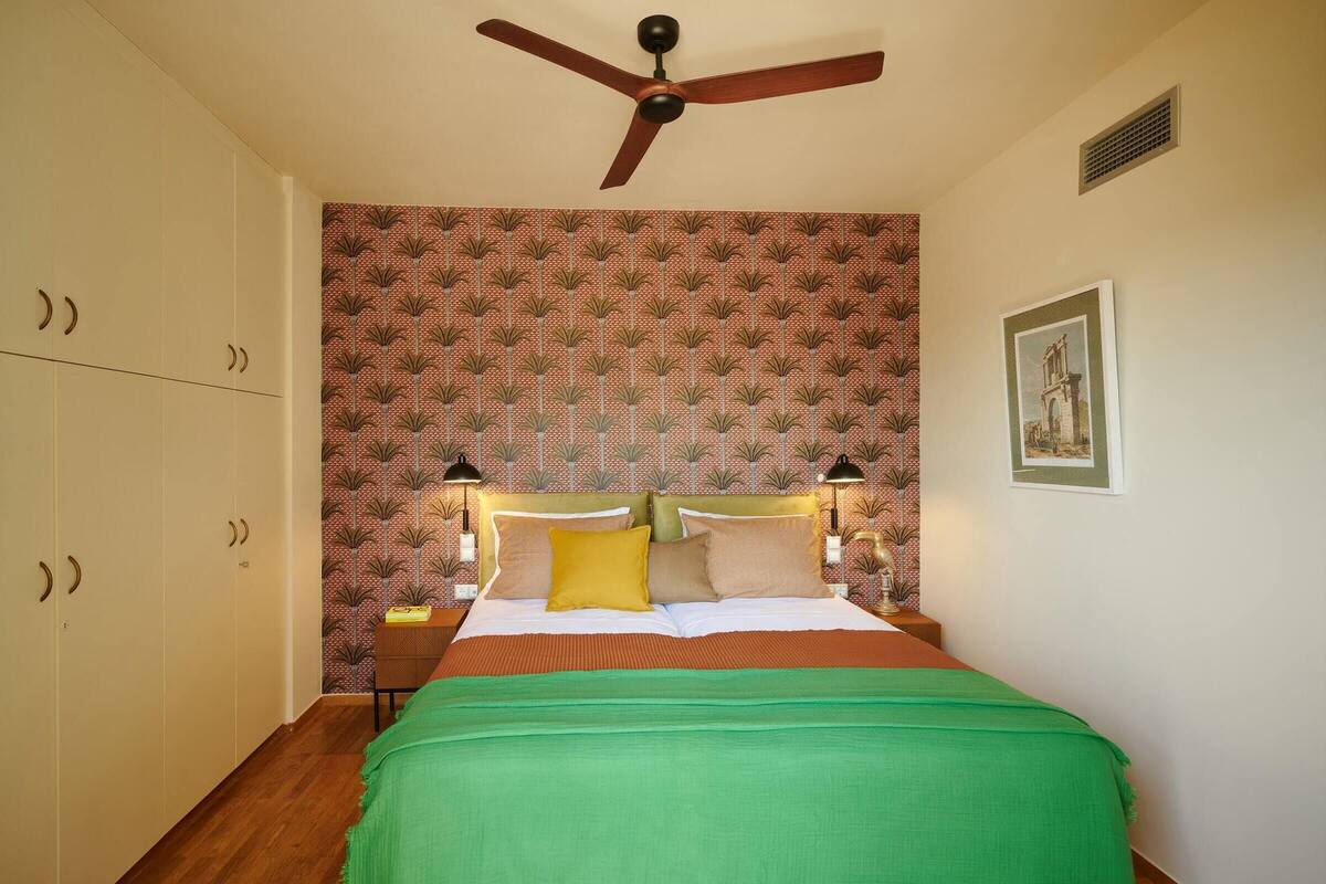 Ein Schlafzimmer mit Bett, Deckenventilator, gemustertem Tapeten und einem gerahmten Bild an der Wand.