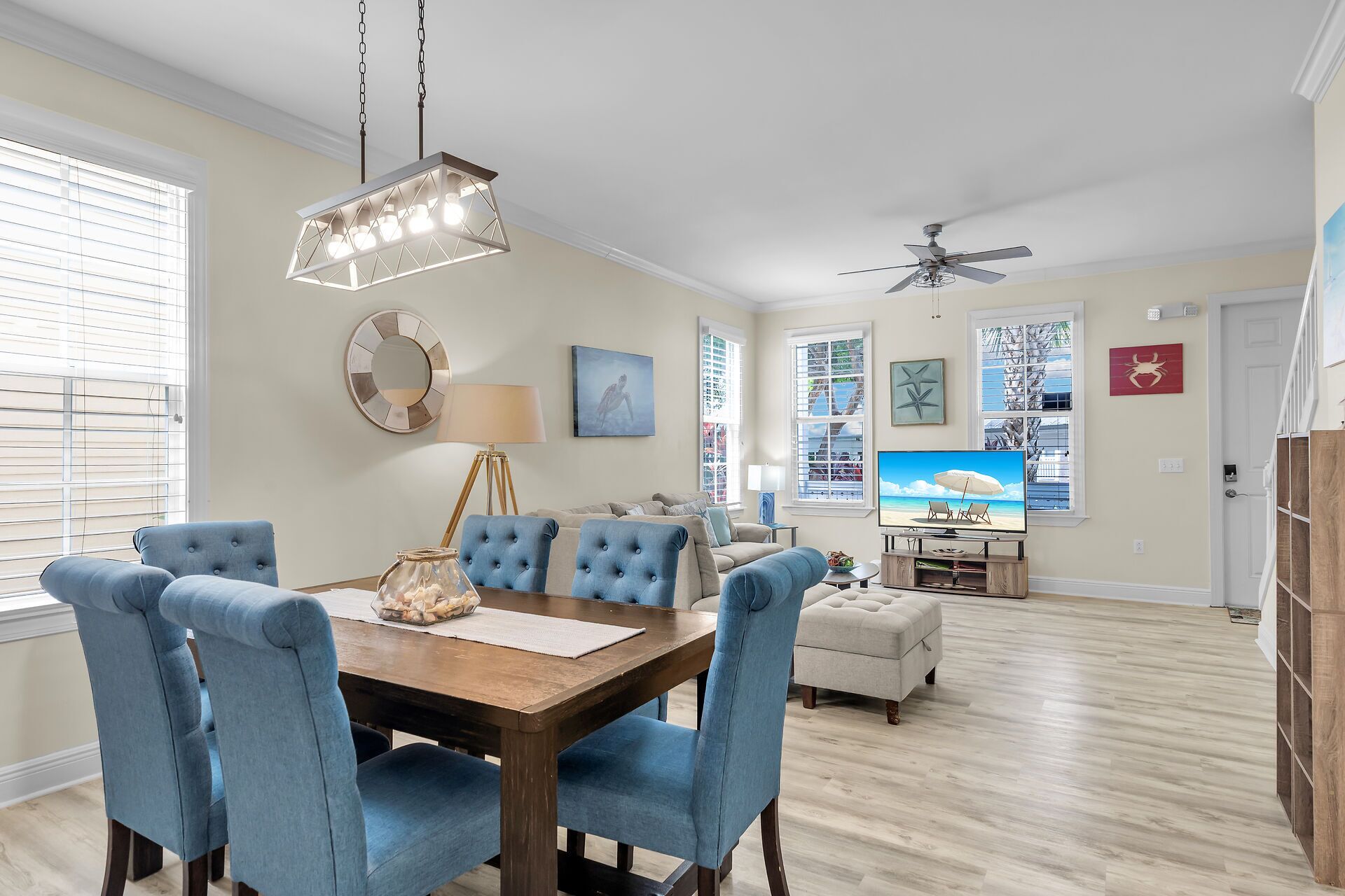 Un coin repas avec une table en bois et des chaises recouvertes d’un tissu bleu, un ventilateur de plafond et une vue sur la plage grâce à de grandes fenêtres.