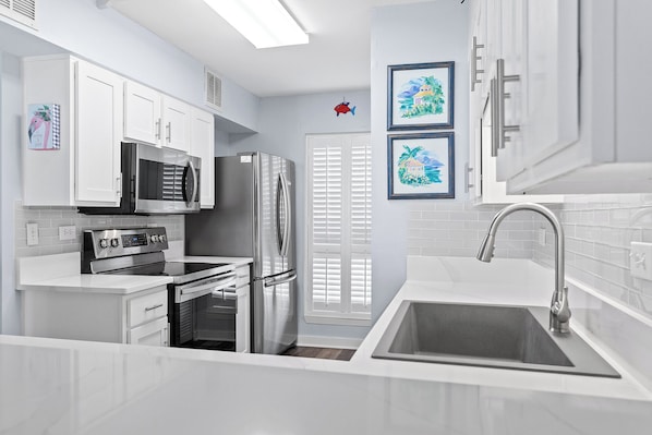 A modern kitchen with white cabinetry, a stainless steel refrigerator, a microwave, and an oven. There are two framed pictures on the wall and a red fish decoration on the door. - Mainsail 352 - Gulf Front Condo, Community Pool, Hot Tub, Free Beach Service! (Miramar Beach)