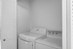 A laundry room with a front-loading washer and dryer, a door with horizontal louvered panels, and a control panel with various settings. - Mainsail 352 - Gulf Front Condo, Community Pool, Hot Tub, Free Beach Service! (Miramar Beach)