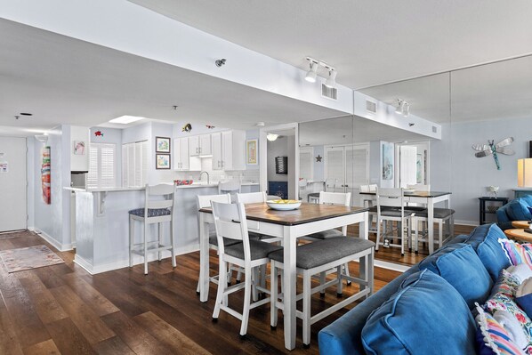 A modern kitchen with white cabinets, a dining area with white chairs, and a blue sofa in the living room. - Mainsail 352 - Gulf Front Condo, Community Pool, Hot Tub, Free Beach Service! (Miramar Beach)