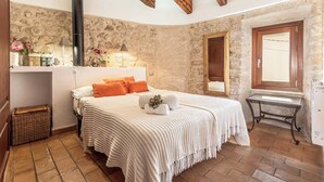 A cozy bedroom with a stone wall, a wooden ceiling, a bed with white bedding and orange pillows, a bedside table with a lamp, and a small wicker basket.