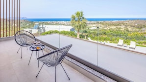 Un balcon avec deux chaises et une table, donnant sur un paysage côtier avec un palmier et des bâtiments situés au loin.