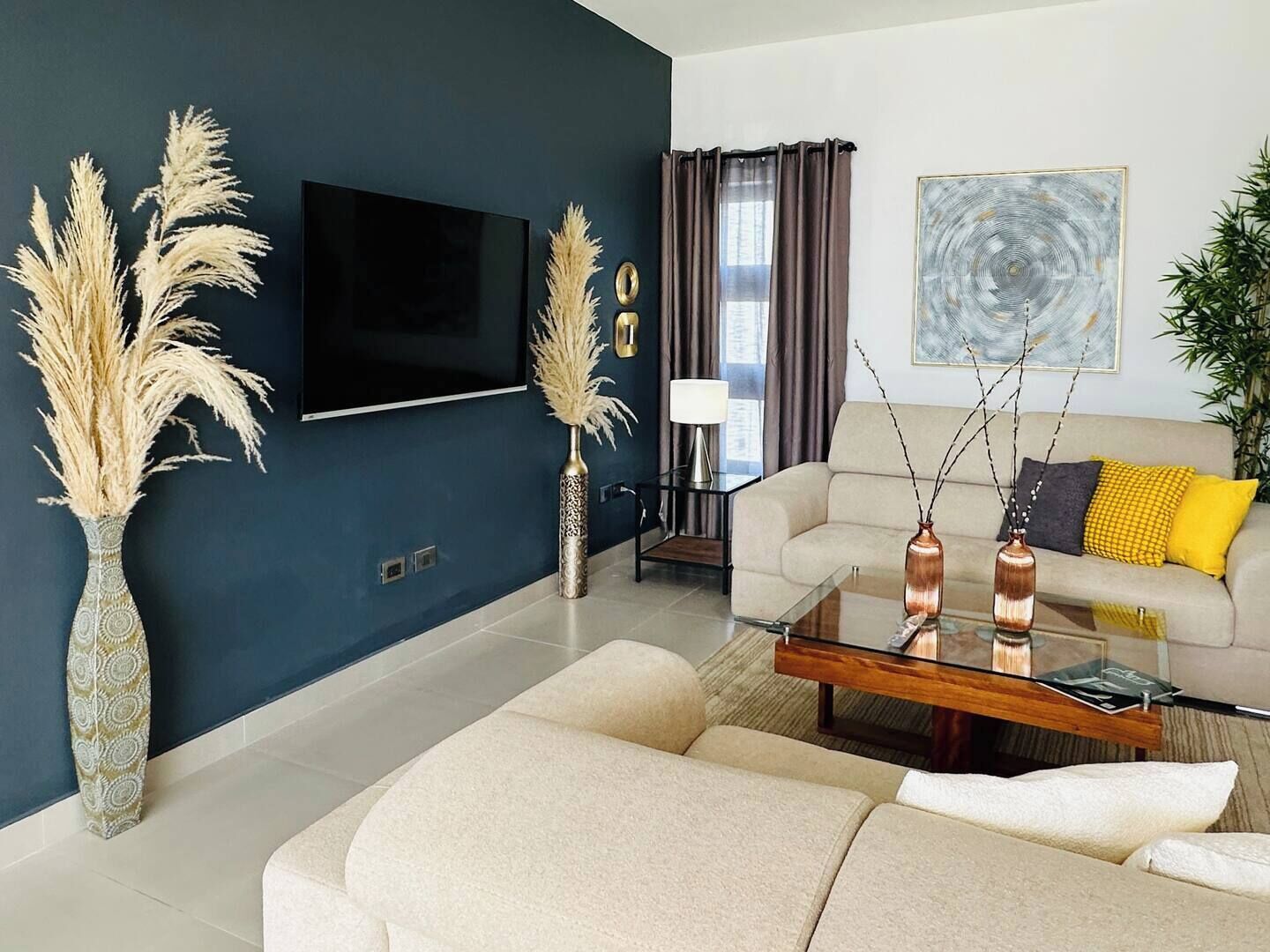 Ein modernes Wohnzimmer mit dunkelblauer Akzentwand, einem Flachbildfernseher, einem beigefarbenen Sofa mit gelben Kissen, einem Glastisch und dekorativen Vasen mit Pampasgras.