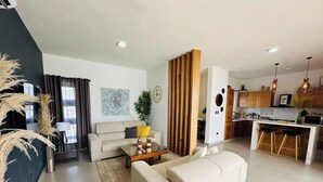 A modern living room with a sofa, coffee table, and decorative plants. A kitchen area with bar stools and a dining table is visible in the background. - Coastal Luxe SOV: 3 Bed, 3.5 Bath, Sosúa Ocean Village (Sosúa)