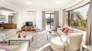 Un salon moderne avec un canapé, une table basse et un téléviseur. Il y a un balcon avec des rideaux et une vue sur la verdure.