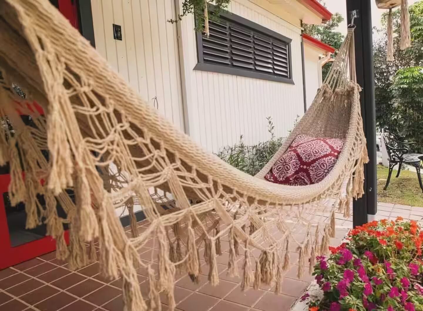 A hammock on a porch with a patterned cushion, a white building with a window and shutters, and a garden with flowers.
