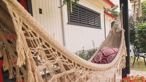 A hammock on a porch with a patterned cushion, a white building with a window and shutters, and a garden with flowers. - Home with Pool & Pickleball Court near El Yunque (Palma Sola)