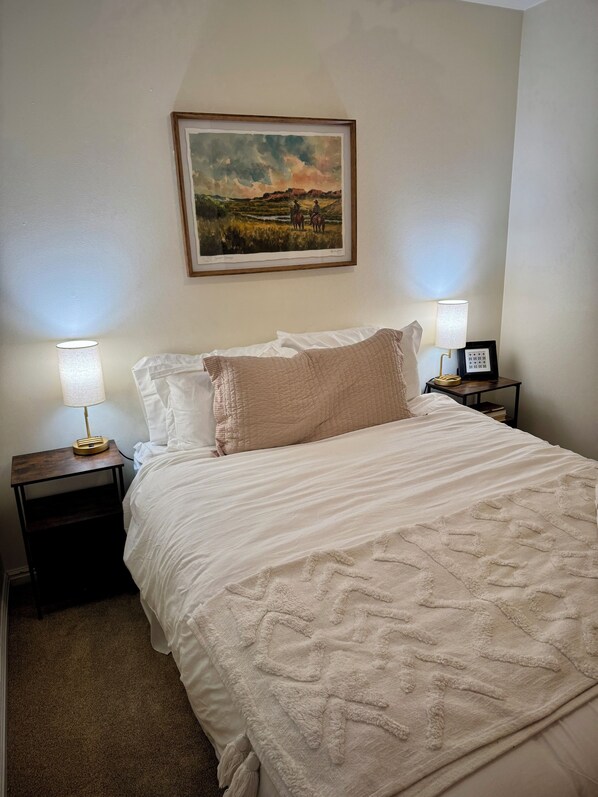 A neatly made bed with a white comforter and a beige throw blanket, flanked by two bedside tables with lamps and framed artwork.