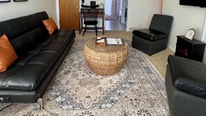 Un salon avec un canapé en cuir noir, une table basse ronde en fibres tressées et un tapis à motifs. Un téléviseur est fixé au mur, et des tableaux encadrés y sont accrochés.