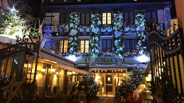 Ein Hotelgebäude mit Weihnachtsdekorationen, darunter beleuchtete Weihnachtsbäume und Ornamente, und eine Schilder mit dem Schriftzug "MARIA & JOHANN".