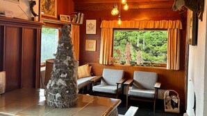 Ein gemütliches Blockhaus-Interieur mit Holzwänden, Kamin, Weihnachtstannenbaum und großem Fenster mit Blick auf Grün.