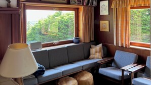 Ein gemütliches Blockhaushaus-Interieur mit Holzbalkendecke, einem großen Fenster mit Vorhängen, einer Couch, Poufs und einer Lampe.