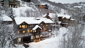 A large, multi-story wooden house with a covered porch and a garage, surrounded by snow-covered trees and hills.