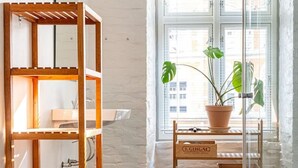 A corner of a room with a wooden shelf unit, a red plastic basket, a wooden desk with a potted plant, and a window with blinds.
