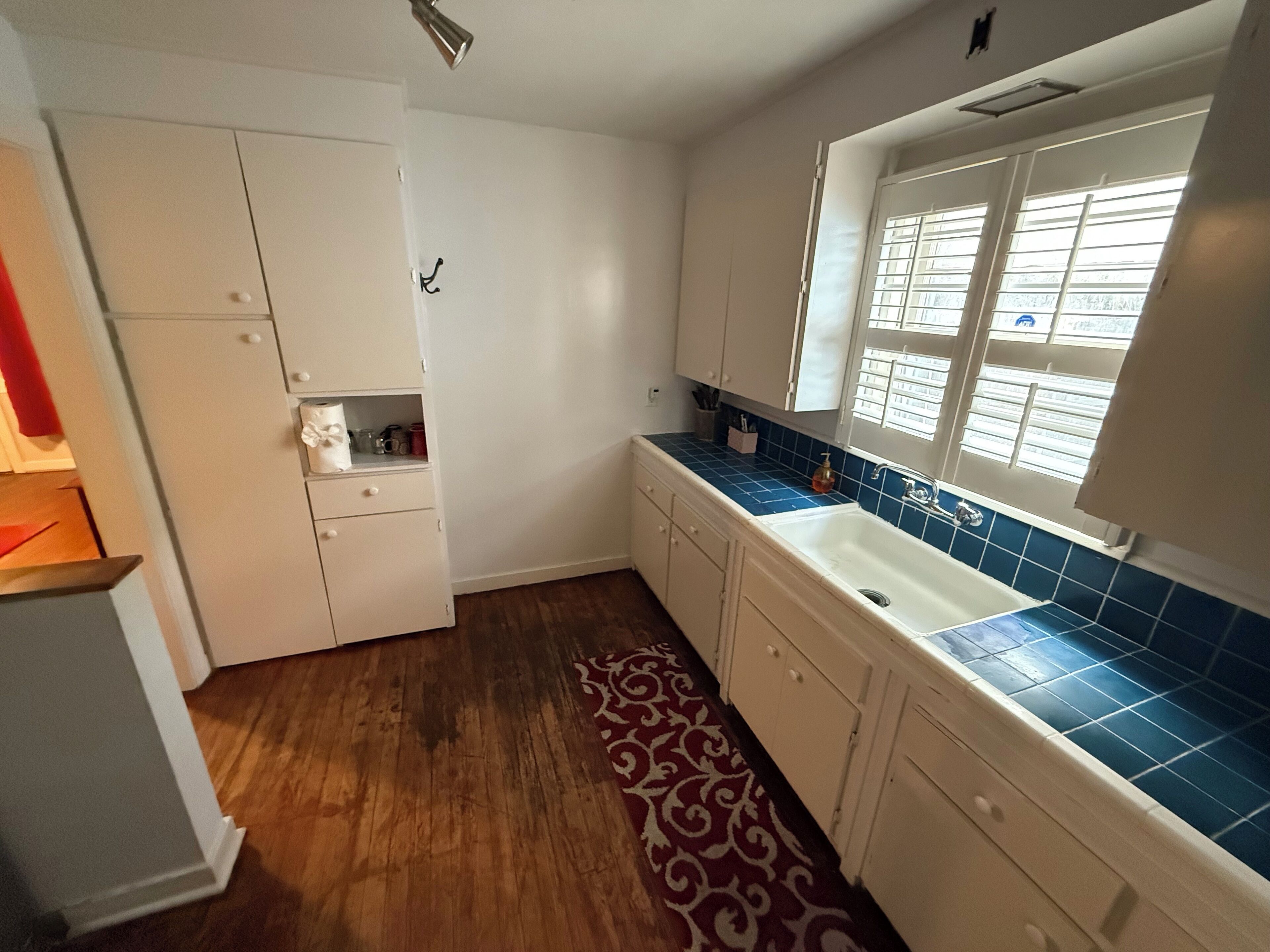A kitchen with white cabinets, a blue tiled backsplash, a window with blinds, and a patterned rug.