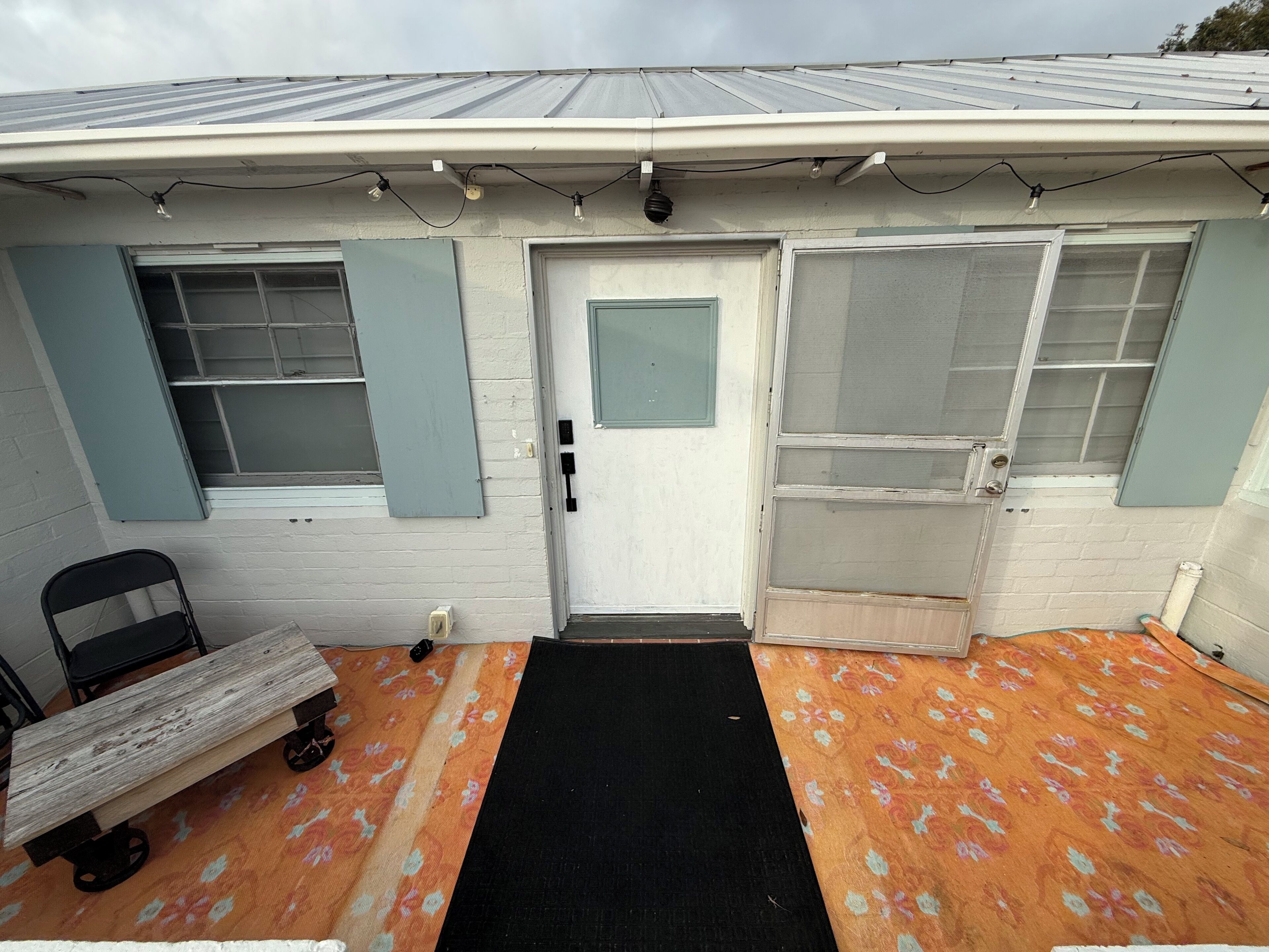 A small house with a front door, two windows with shutters, a bench, and a black mat leading to the door.