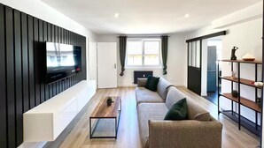 Uma sala de estar moderna com uma televisão de tela plana fixada em um painel preto, uma mesa de centro de madeira, um sofá bege com almofadas verdes e um piso de madeira.