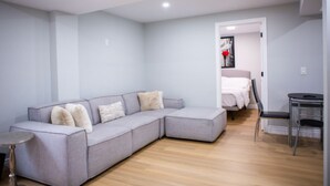 A modern living room with a grey sectional sofa, a small round table, and a bed in the background. - Cozy and Private Basement apartment / Ideal for Relaxation or Work (Brampton)