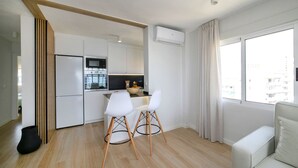 Cozinha moderna com armários brancos, uma geladeira preta e um micro-ondas. Há uma área de jantar com duas cadeiras brancas e uma mesa. Uma janela grande oferece vista para a cidade.