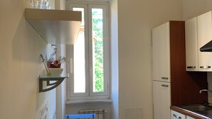 A compact kitchen with white cabinets, a window with a view of greenery, a table with a citrus-patterned tablecloth, and a hanging light fixture. - Flat nearby Vaticano - Caracciolo 4 (Rome)