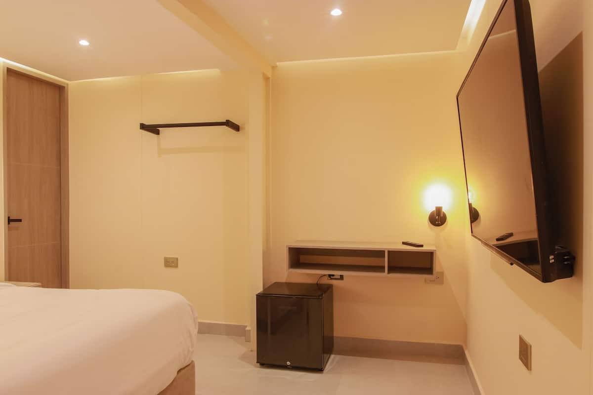 A hotel room with a bed, a small desk, a mirror, and a shelf with a light.