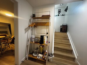 Et kompakt køkken med kaffemaskine, hylder og et spiseområde med bord og stole.