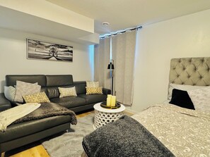 Et moderne soveværelse med en grå sofa, en seng med en polstret gavl, et sengebord med en lampe og et billede på væggen.