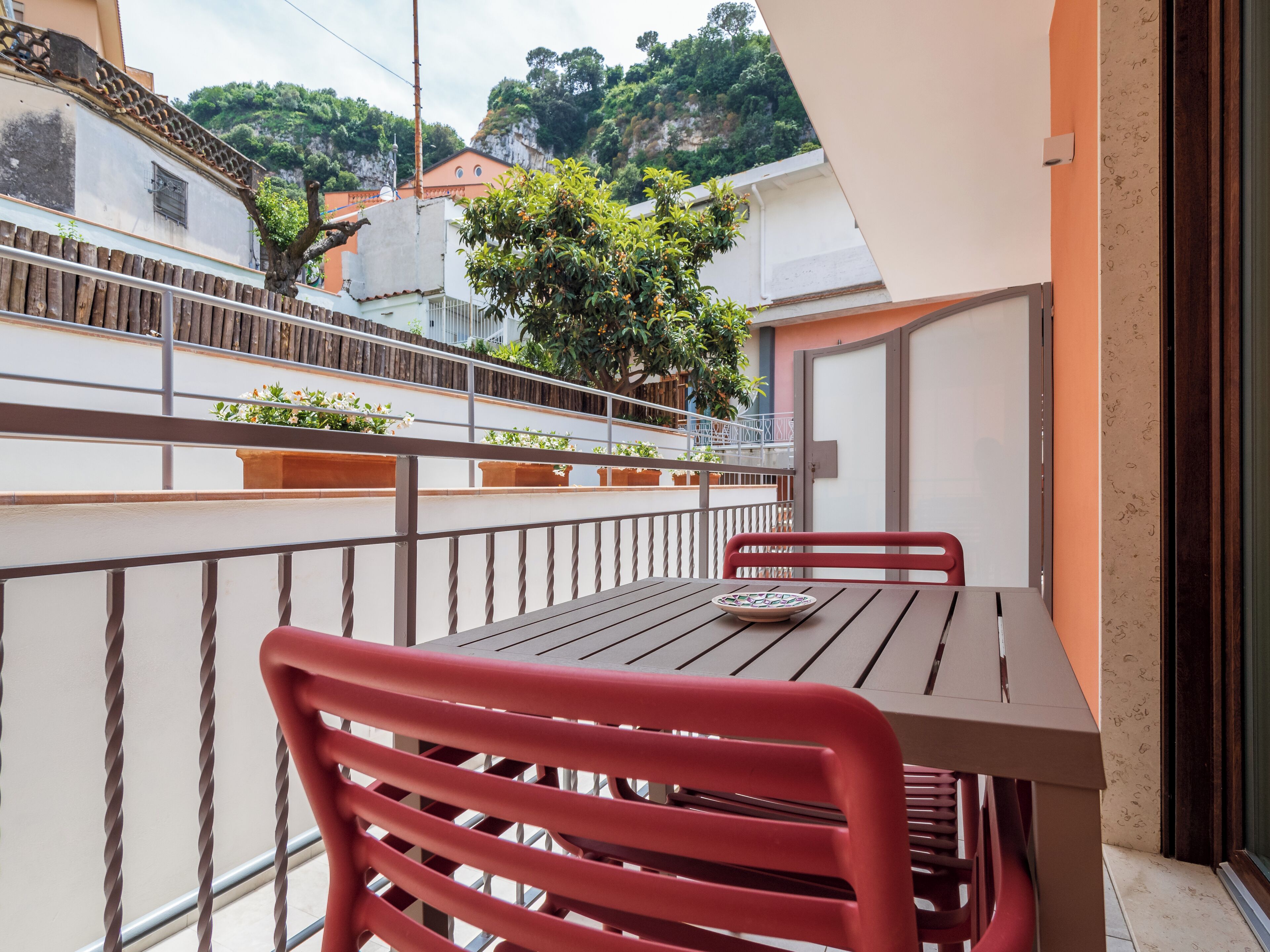 Ein Balkon mit Tisch und roten Stühlen, mit Blick auf eine Wohngegend mit Häusern und Grünflächen.